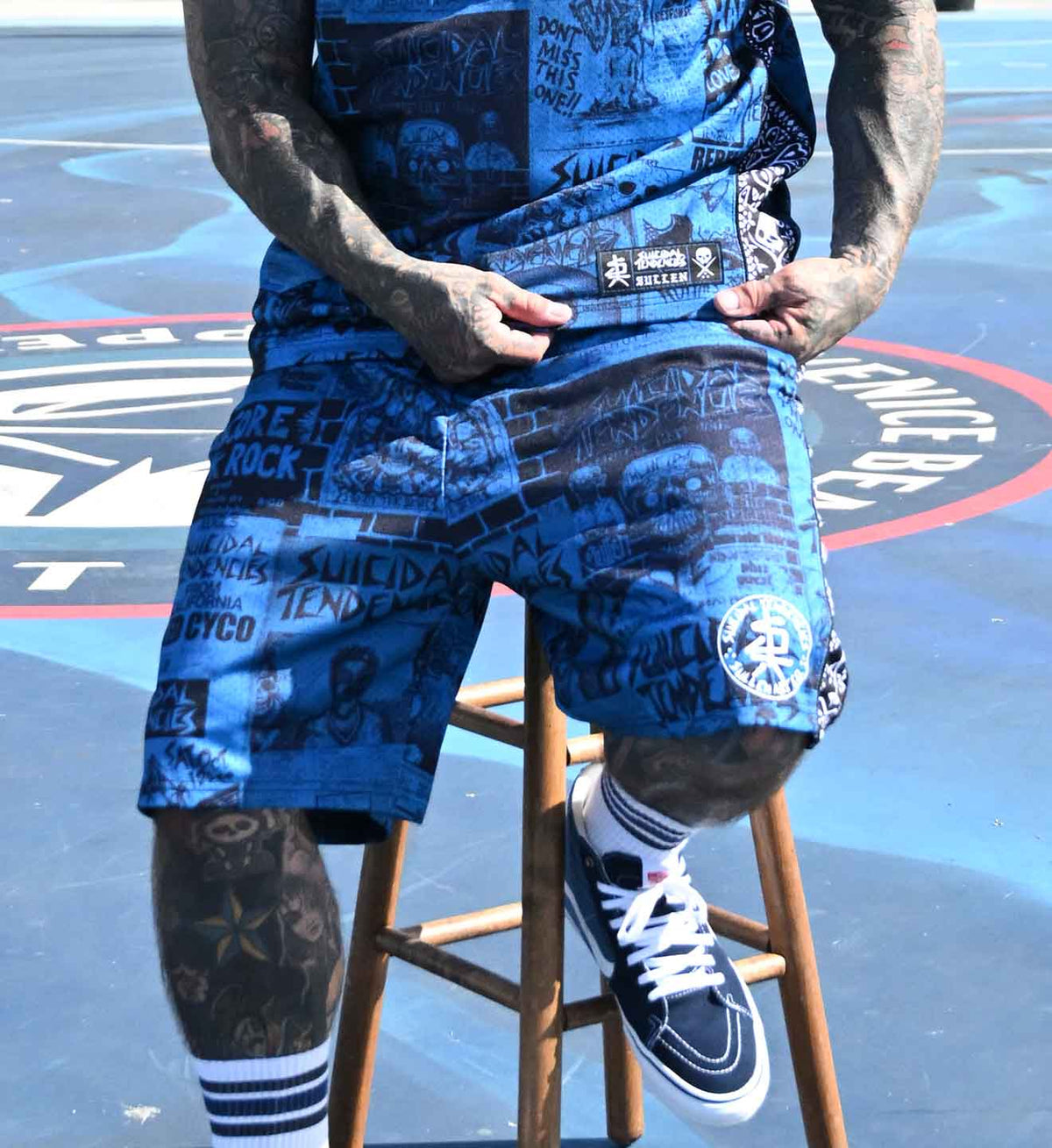 A person seated on a wooden stool wearing blue graphic shorts and a matching blue sleeveless top, with tattooed legs and white-and-black sneakers, photographed on a painted outdoor court.