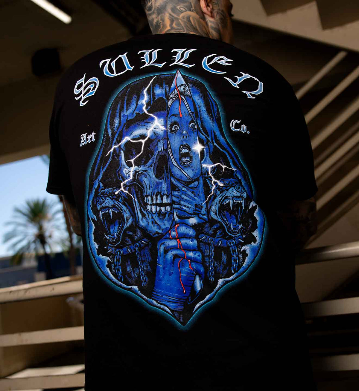 A man viewed from behind wearing a black t-shirt with a large graphic print on the back. The graphic features a blue-toned skull, roses, lightning, and the text 'SULLEN Art Co.