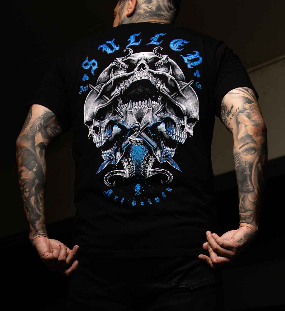 A man wearing a black t-shirt from the back, featuring a large graphic print of three skulls with crossed knives and blue gothic text reading 'SULLEN ART DRIVEN'.