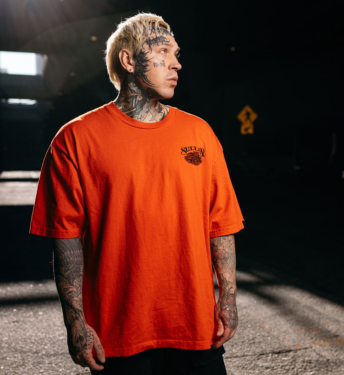 Man wearing an orange t-shirt with a logo, standing in a dark outdoor setting.