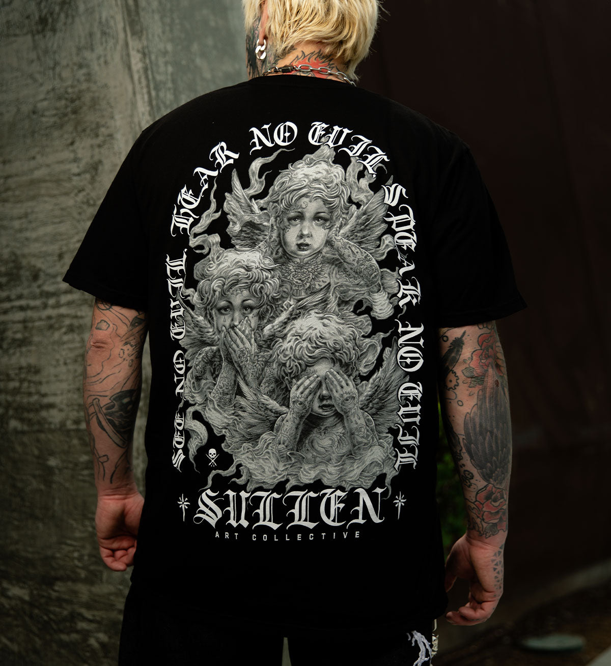 Person wearing a black t-shirt with a graphic design and text on the back, standing against a dark background.