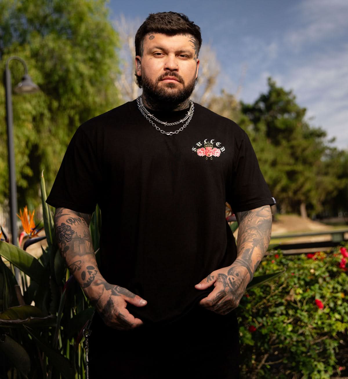 Man wearing a black t-shirt with a logo, standing outdoors with greenery in the background