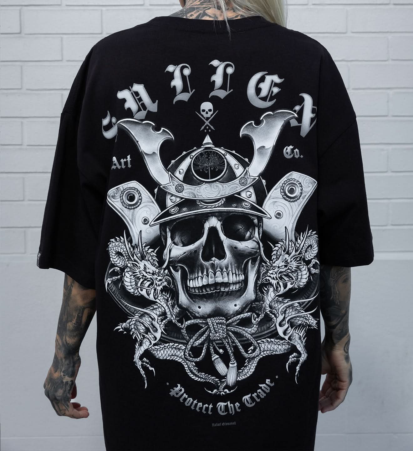 Person wearing a black t-shirt with a detailed skull and crossbones design on the back, against a white brick wall.