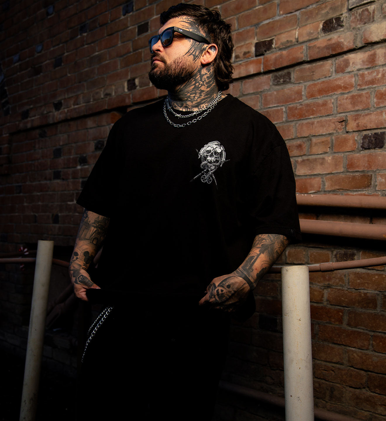 Man with tattoos wearing a black t-shirt with a graphic design, standing against a brick wall.