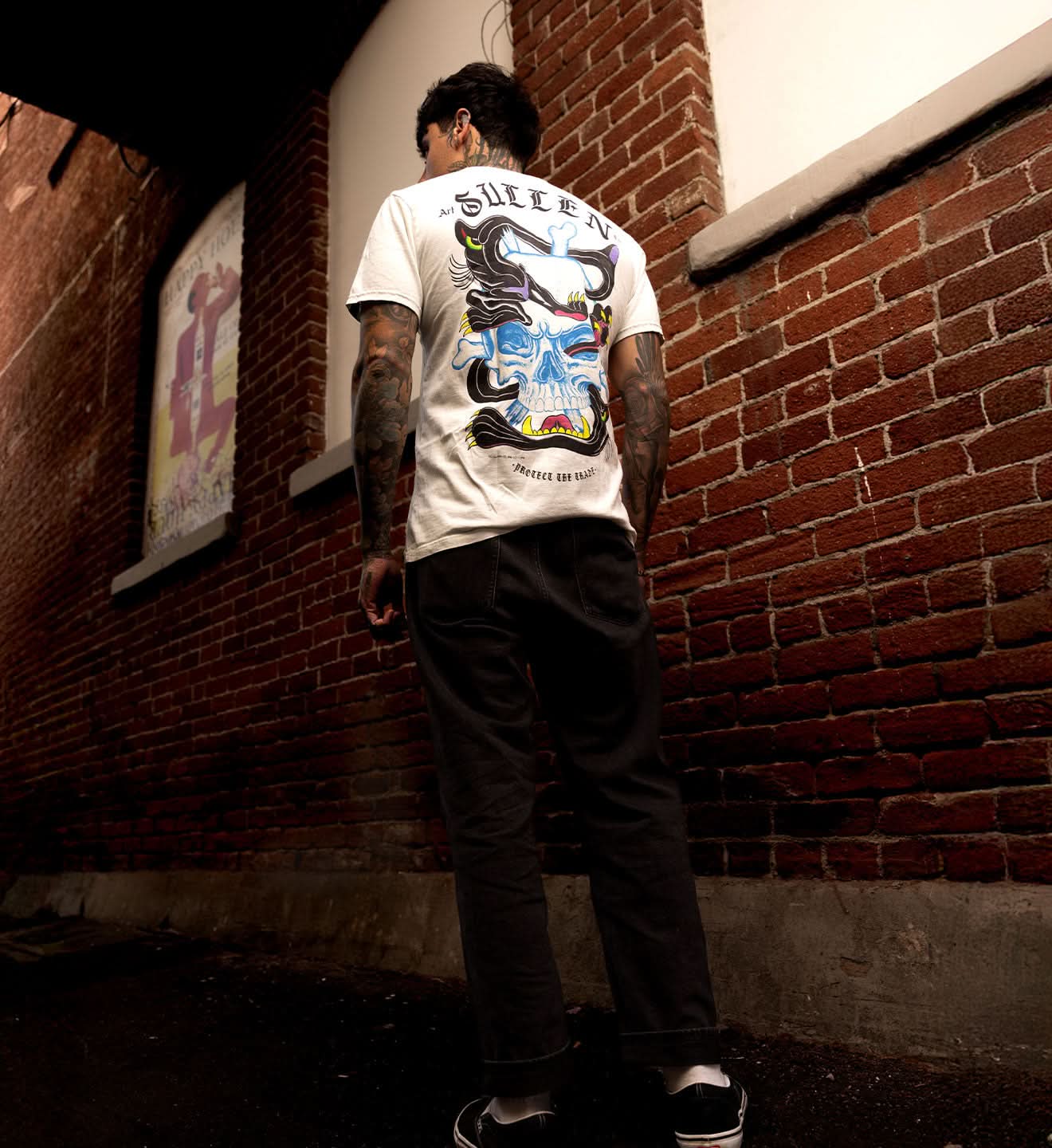 Person wearing a white t-shirt with a colorful design, standing against a brick wall.