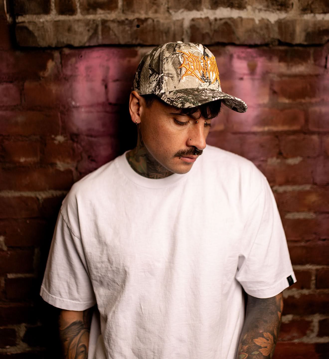 Man wearing a camouflage cap with a logo against a brick wall