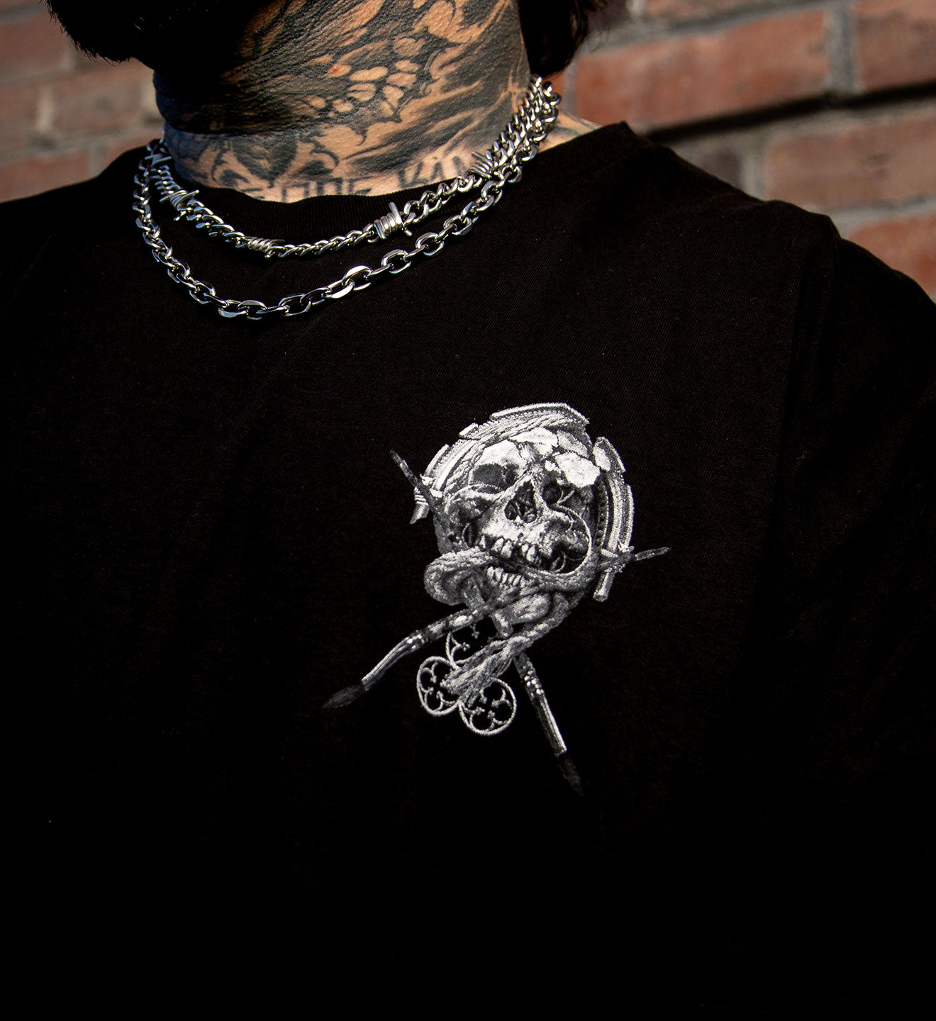 Person wearing a black shirt with a skull and snake design, standing against a brick wall.
