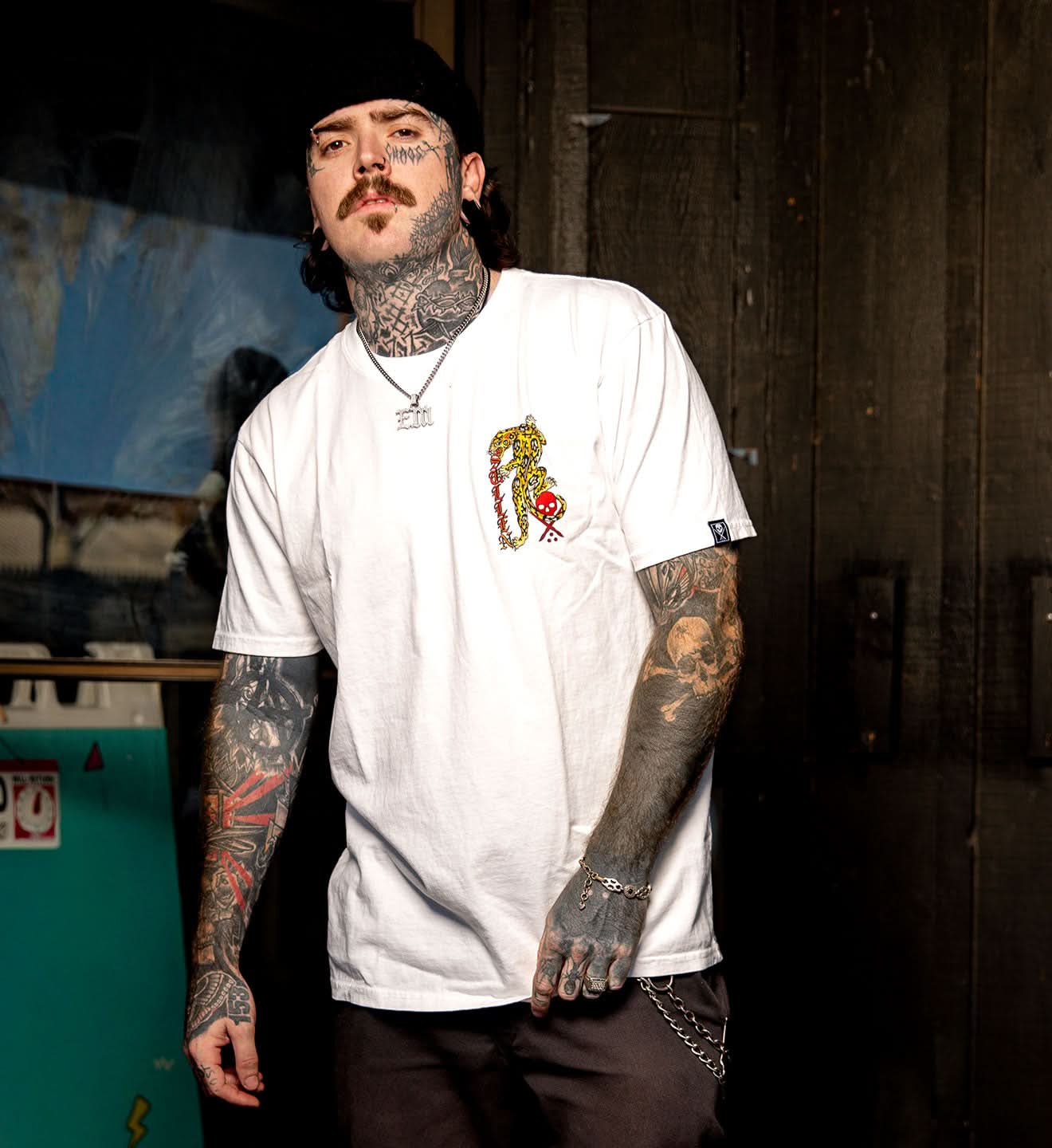 Man with tattoos wearing a white t-shirt with a colorful graphic design in a dimly lit room.