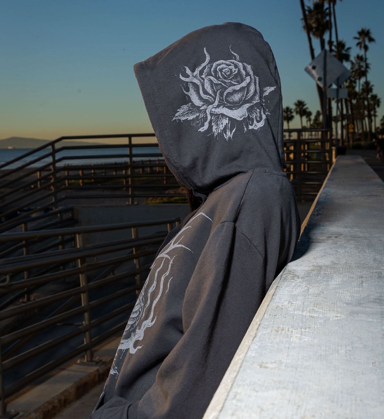 Person wearing a dark hoodie with a rose design, standing on a pier with palm trees in the background.