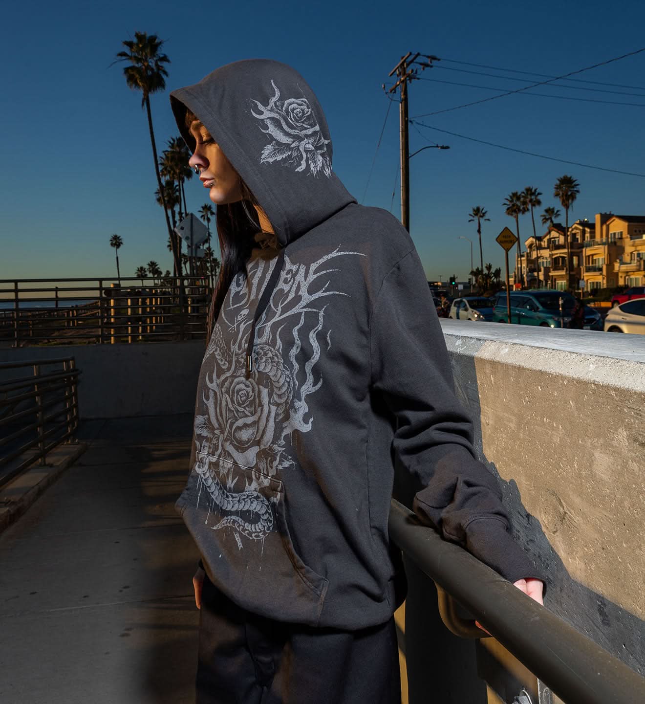 Person wearing a dark hoodie with a white design, standing on a pier with palm trees and buildings in the background.