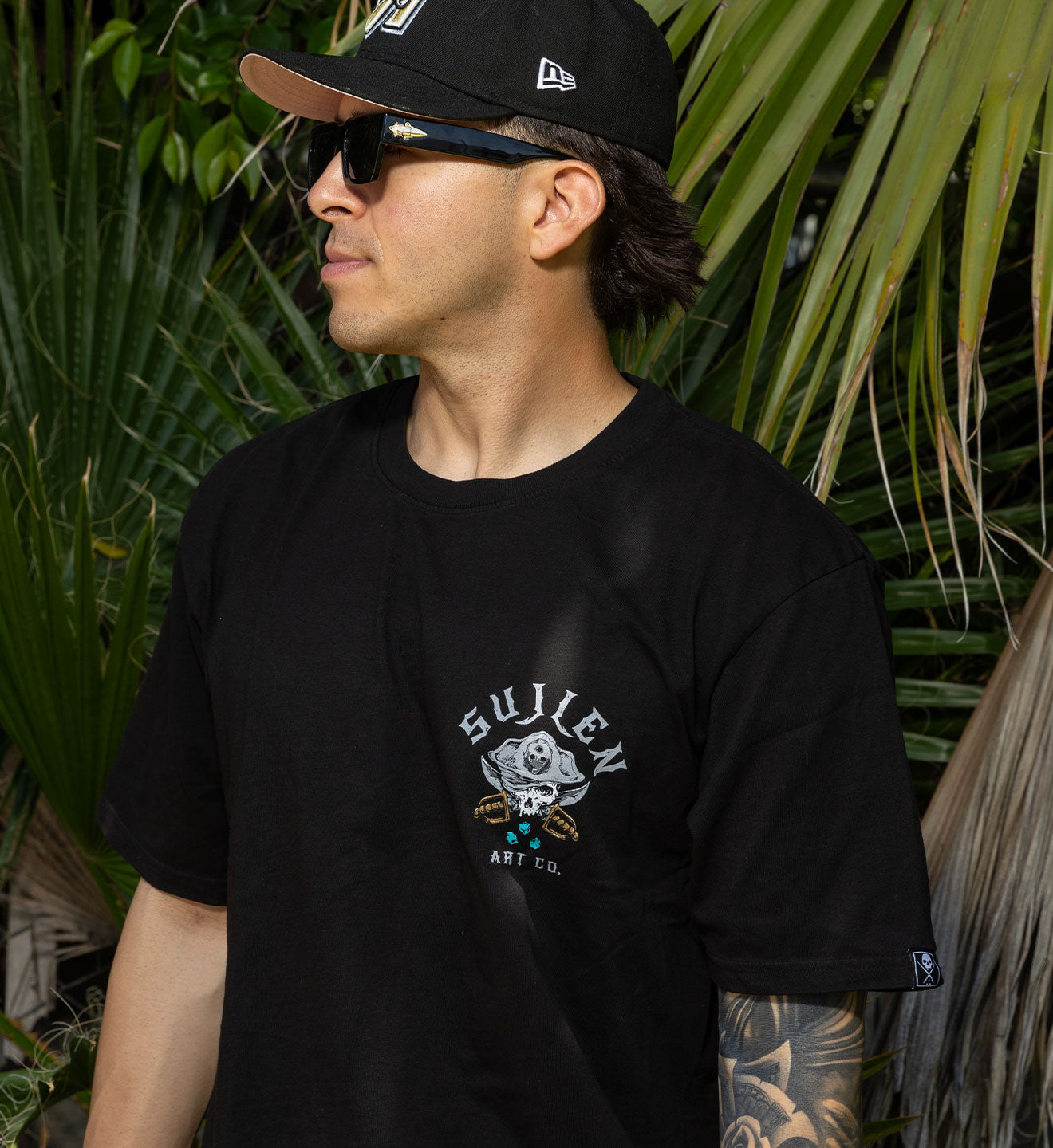 Man wearing a black t-shirt with a logo and text, sunglasses, and a cap in front of green foliage.