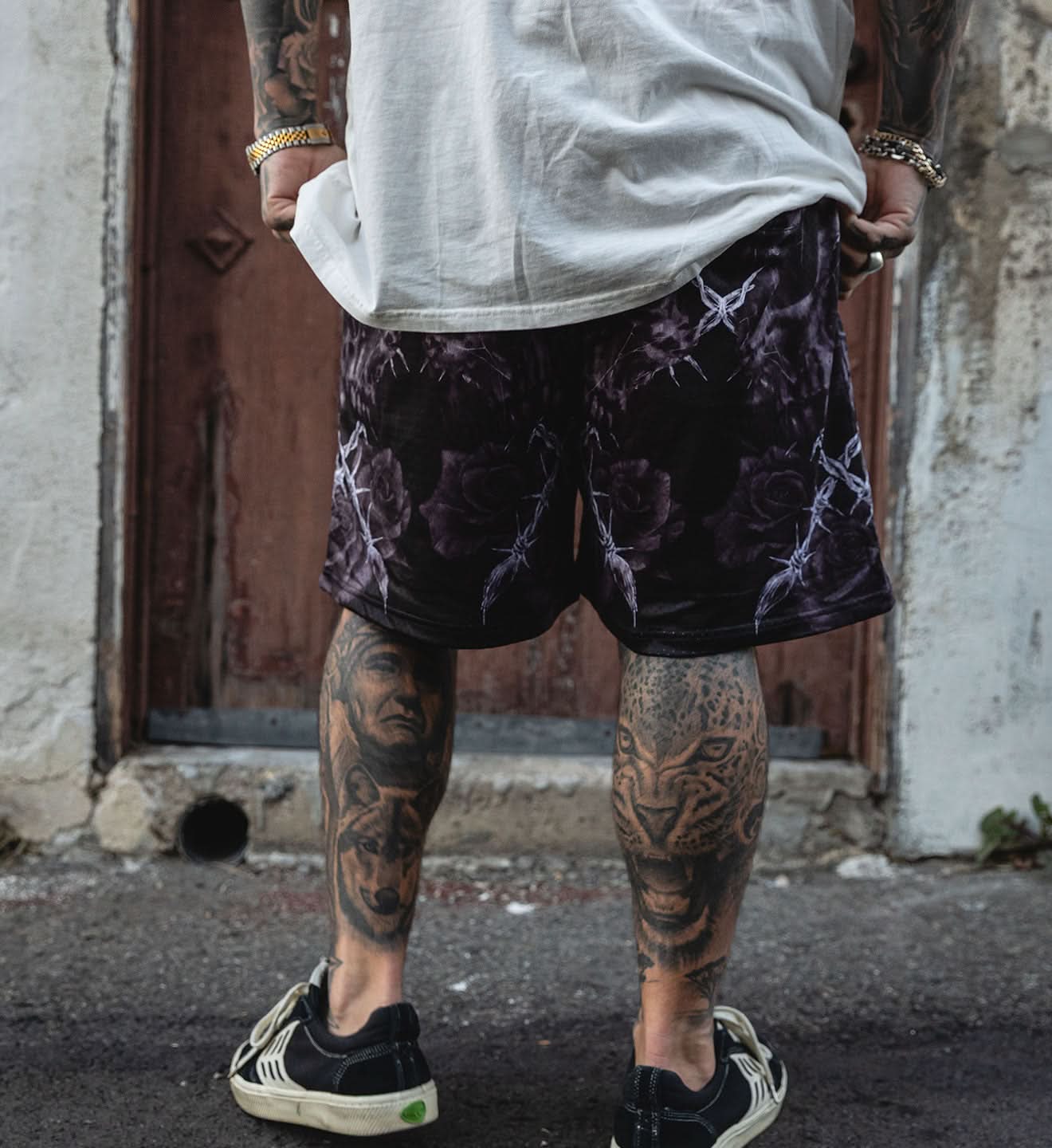 Person wearing black shorts with purple patterns and tattoos on legs, standing against a textured wall.
