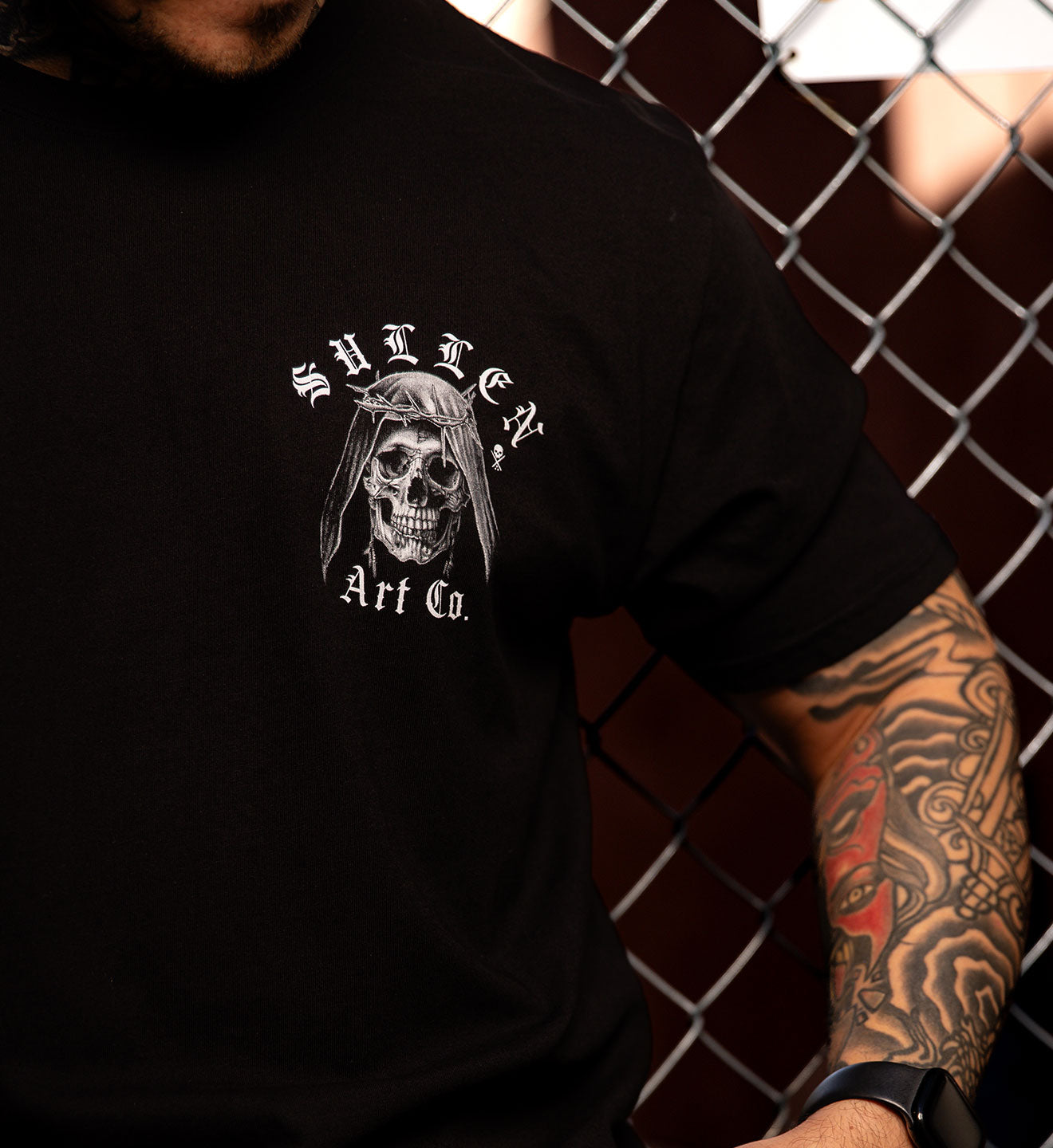 Person wearing a black t-shirt with a skull design and text, standing in front of a chain-link fence.